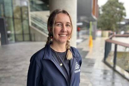 A smiling woman with fair skin, green eyes, and light blonde hair styled in two braids. She has a friendly, open expression with a wide smile and a subtle nose stud. She is wearing a navy blue zip-up jacket over a dark t-shirt and a thin gold chain necklace with a small circular pendant. Her earrings are small circular dangles featuring the black, red, and yellow colors of the Australian Aboriginal Flag.
