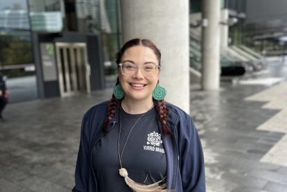A smiling woman with reddish-brown hair styled in two long braids. She wears clear-rimmed glasses, large circular green woven earrings, and a long cord necklace featuring a pendant with large, light-colored feathers. She is dressed in a dark navy blue zip-up jacket over a black t-shirt.